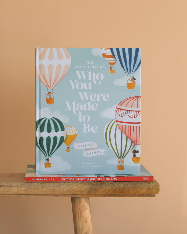 The World Needs Who You Were Made To Be," a children’s book celebrating creativity, features colorful hot air balloons on the cover and is displayed upright against a peach background.
