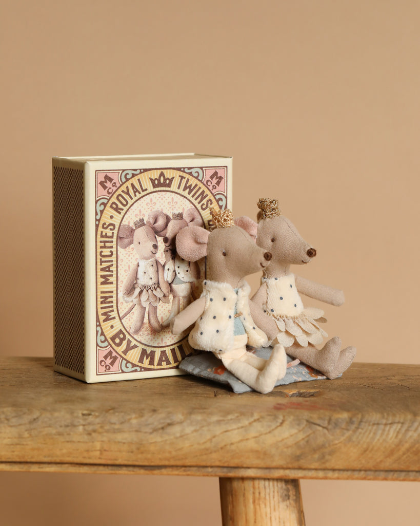 Two Maileg Royal Twin Mice, Little Sister and Little Brother, wearing white outfits, sit on a wooden surface leaning against a large decorated matchbox labeled "Royal Twins by Maileg," set against a warm, neutral background.