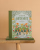 We Are The Gardeners," a children's book, stands upright on a wooden bench with another copy below it against a beige background. The cover features illustrated kids learning from their family's thriving garden.