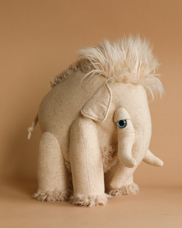 A cream-colored BigStuffed Albino Mammoth Stuffed Animal stands against a plain tan background. Featuring a fluffy mane and tail, with a single blue eye visible, this whimsical mammoth design emphasizes its soft and cuddly appeal.