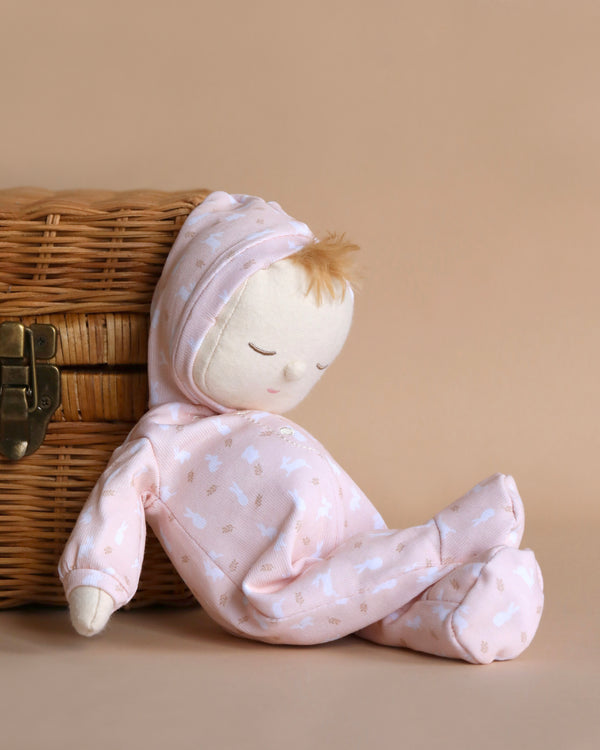 A Dozy Dinkums - Blossom from the Olli Ella x Odin Parker collection with a serene face, wearing a pink outfit and matching hat, leans against a wicker basket on a beige background.
