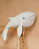 The BigStuffed The Big Whale Albino Stuffed Animal, with its textured fabric and blue eye, drapes over a wooden ladder against a beige backdrop. Ideal for imaginary quests, it features a soft and cuddly design with a light-colored underside and slightly darker upper side.