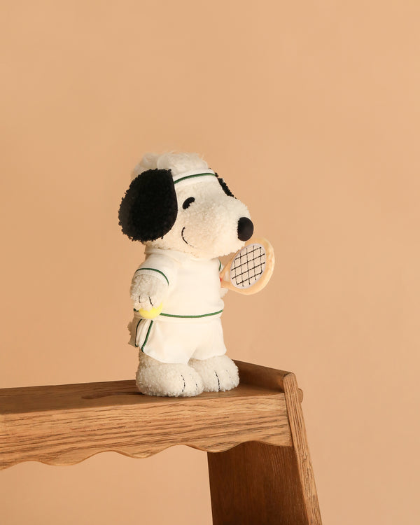 The Plush Peanuts Snoopy Tennis stands on a wooden stool, wearing a white tennis outfit and holding a racket and yellow ball, set against a plain beige background.