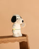 The Plush Peanuts Snoopy Tennis stands on a wooden stool, wearing a white tennis outfit and holding a racket and yellow ball, set against a plain beige background.
