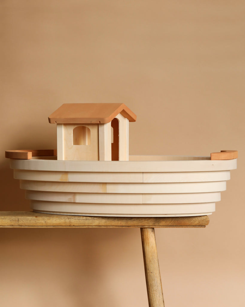 The Holztiger Handmade Wooden Ark, crafted from light maple and beech wood, sits on a wooden bench. Its simple, layered design against a beige backdrop enhances any HOLZTIGER figure collection with its natural charm.