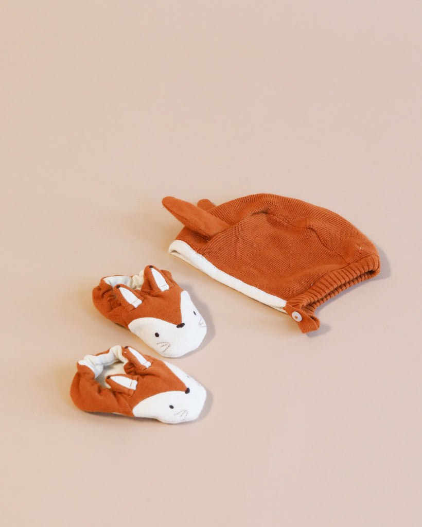 A pair of small, Meri Meri Fox Bonnet & Booties Set and a matching terracotta-colored organic cotton bonnet arranged neatly on a soft pink background.