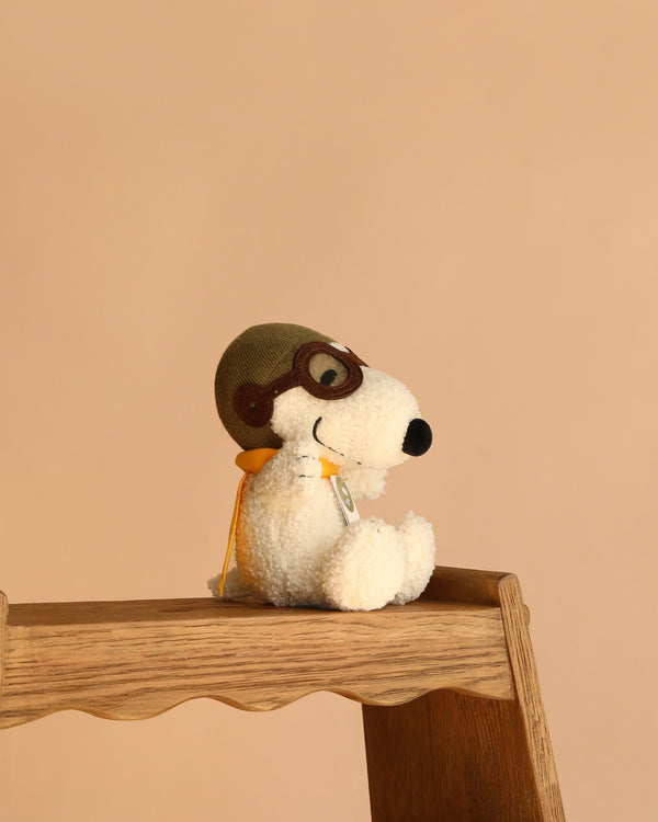 The Plush Peanuts Snoopy Sitting Flying Ace wears aviator goggles, a green cap, and a yellow scarf while sitting on a wooden step stool against a plain beige background.