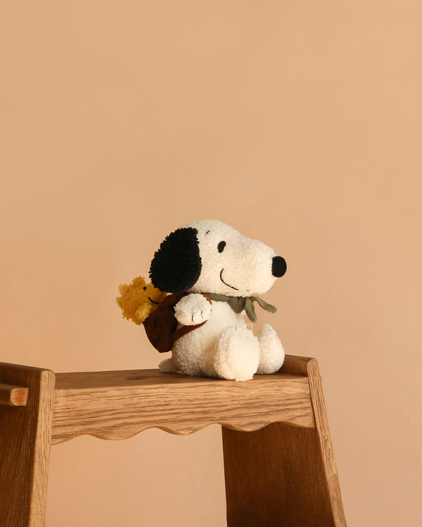 The Plush Peanuts Snoopy with Woodstock in Backpack, featuring black ears and a brown backpack, sits on a wooden step stool against a plain beige background.