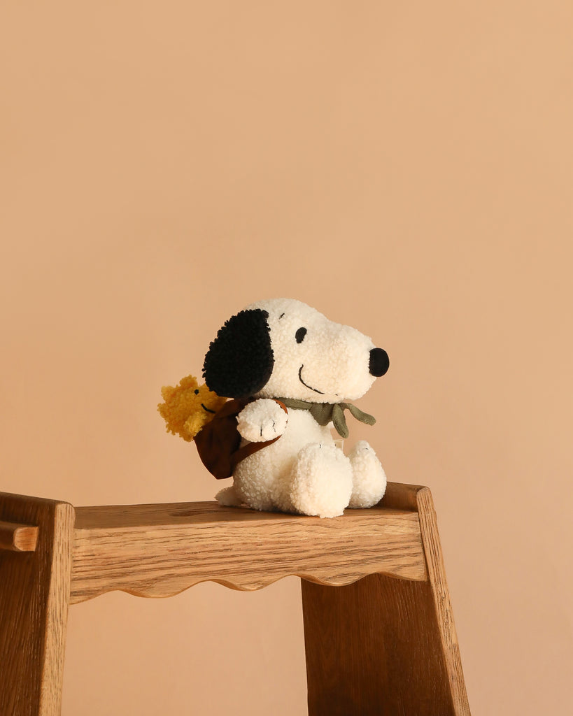The Plush Peanuts Snoopy with Woodstock in Backpack, featuring black ears and a brown backpack, sits on a wooden step stool against a plain beige background.