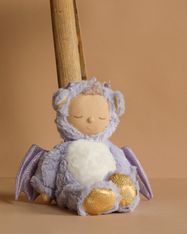 Olli Ella Cozy Dinkum Dragon - Rory - Lavender sits against a peach backdrop, wearing a winged costume with gold glittery feet and closed embroidered eyes, ready to offer a calming cuddle for little ones.