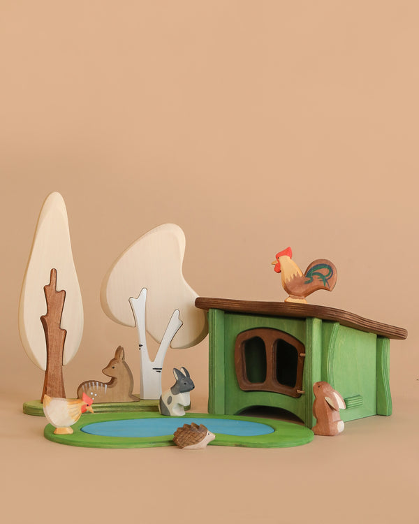 The Enchanted Woodland Farm Set includes handmade wooden animals—a rooster, duck, rabbit, hedgehog, deer, and fox—plus Brin d’Ours trees, all arranged around a green toy house and blue pond on a beige background.