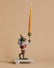 A figurine of a green, anthropomorphic creature with a long nose, wearing brown pants, a red vest, and a cone hat. Crafted from poly resin material, the "Candle Holder - Tiger" stands on a wheeled platform as part of a whimsical birthday train, holding a large tapered yellow candle upright in its right hand against a plain beige background.