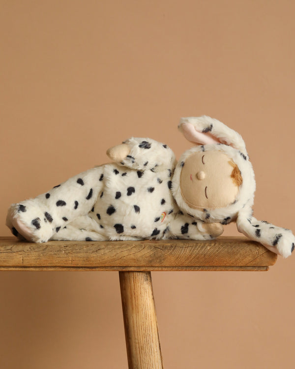 The Olli Ella Cozy Dinkum - Dog Domino plush, wearing a white and black-spotted dog costume with floppy ears, lies on its side atop a wooden stool against a plain beige background.