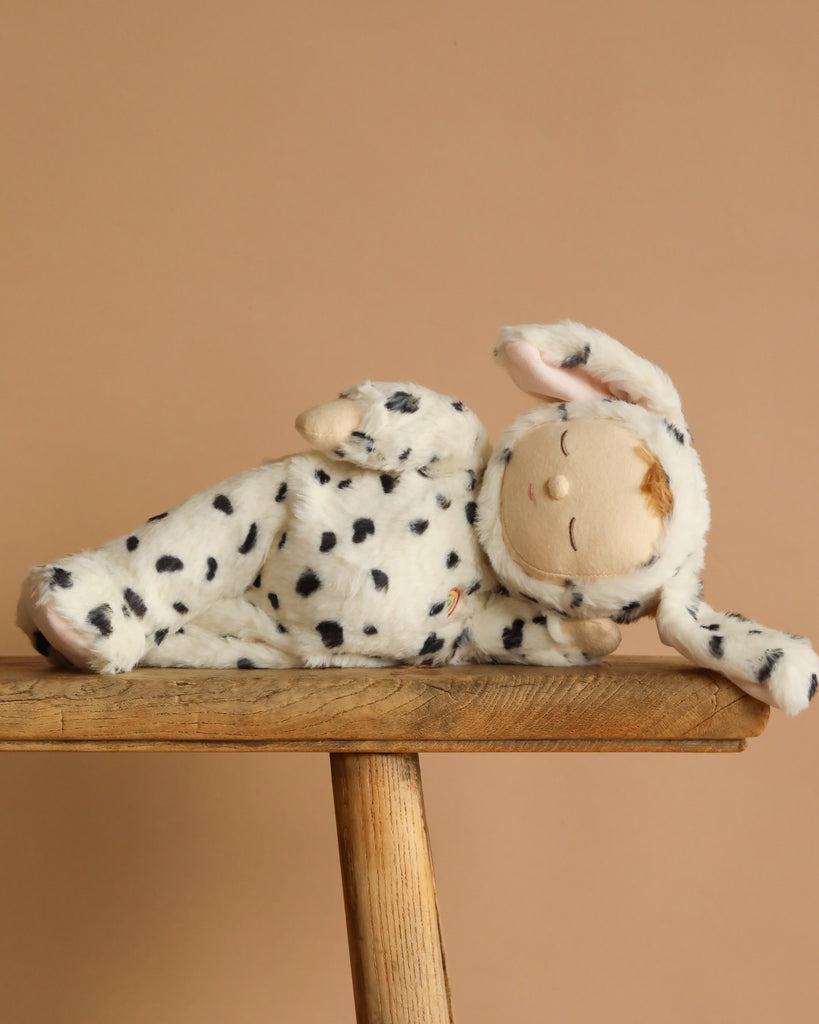 The Olli Ella Cozy Dinkum - Dog Domino plush, wearing a white and black-spotted dog costume with floppy ears, lies on its side atop a wooden stool against a plain beige background.