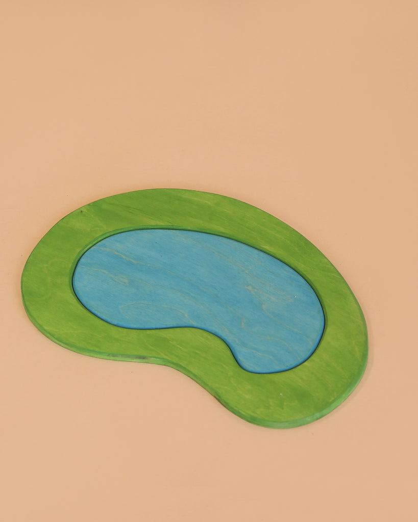 The Enchanted Woodland Farm Set is a flat, bean-shaped handmade wooden toy puzzle with a blue center piece and a green border, displayed on a plain beige background.