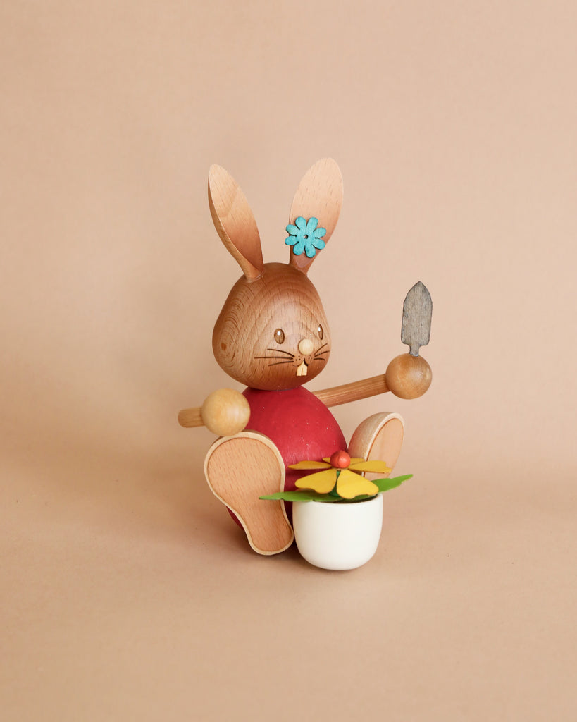 A Collectible Dregeno Easter Figure - Rabbit Gardener With Flower Pot, made in Germany, with a red shirt, holding a trowel and sitting in a white pot filled with colorful flowers. It has a blue flower behind one ear.