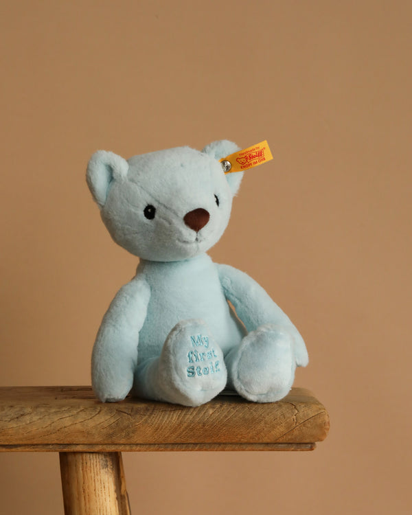 The 10-inch My First Steiff Blue Teddy Bear, featuring "My First Steiff" embroidered on its foot and the signature ear tag, sits on a wooden stool against a beige background—the perfect soft teddy bear gift for babies.