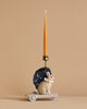 The Candle Holder - Bunny, a delightful addition to a charming birthday train, showcases a small rabbit figurine adorned with a blue cape featuring stars and moons. Made from durable poly resin, the holder rests on a wheeled base, supporting a tall, slender unlit candle that rises from the rabbit's back against a plain beige background.