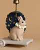 The Candle Holder - Bunny, a charming poly resin figurine of a tan rabbit with a white ruffled collar, sits on a small wheeled platform as part of an enchanting birthday train. Behind the rabbit, there's a navy blue backdrop adorned with gold stars and a crescent moon set against a solid beige background.