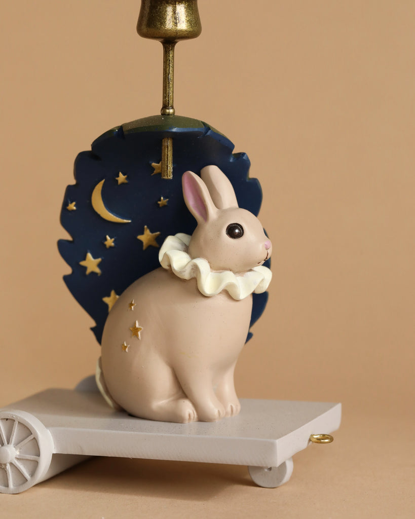 The Candle Holder - Bunny, a charming poly resin figurine of a tan rabbit with a white ruffled collar, sits on a small wheeled platform as part of an enchanting birthday train. Behind the rabbit, there's a navy blue backdrop adorned with gold stars and a crescent moon set against a solid beige background.