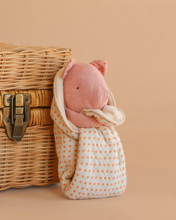 The Maileg Lullaby Friends, Pig, Mini - Rose is a soft pink plush pig wrapped in a cream polka-dotted cuddle cloth, shown beside a wicker basket on a beige background—an ideal gift for newborns.