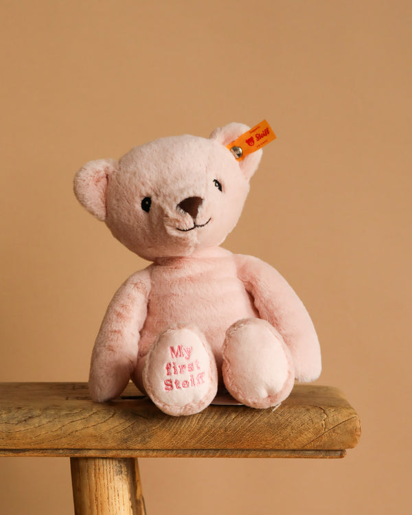 The 10-inch My First Steiff Pink Teddy Bear, featuring a yellow tag and "My first Steiff" pink embroidery on its left foot, sits on a wooden stool against a beige background—an adorable baby gift.