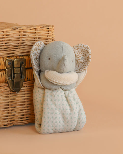 The Maileg Lullaby Friends Elephant, Mini in Iron Grey has patterned ears and a polka-dot body, sitting upright beside a woven basket with a metal latch—a charming newborn gift against a light beige background.