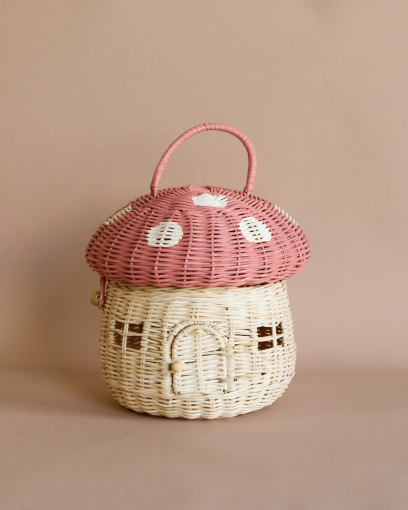 A whimsical handwoven Olli Ella Mushroom Basket - Pink, featuring a pink cap and a white base with a door and windows, set against a soft beige background.