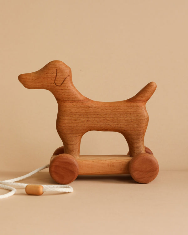 A Handmade Wooden Dog Pull Toy in the shape of a dog is shown against a plain beige background. The toy features four round wheels and a white pull string with a wooden handle, allowing it to be pulled along. The wood grain is visible, giving the Handmade Wooden Dog Pull Toy a natural look.