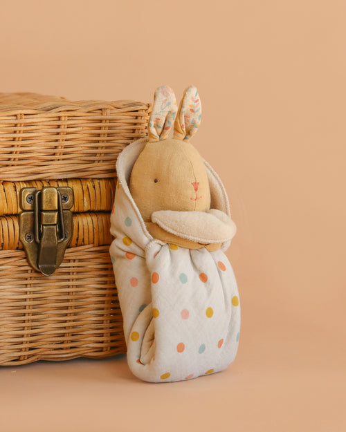 The Maileg Lullaby Friends, Bunny, Mini - Cream Yellow, with floral ears and a polka-dot cuddle cloth, sits by a wicker basket with a metal latch on a beige background.