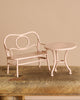 The Maileg Bonus Room Bathroom Set features a small, light pink metal bench and round table on a rustic wooden surface—perfect miniature dollhouse accessories—displayed against a plain beige background.