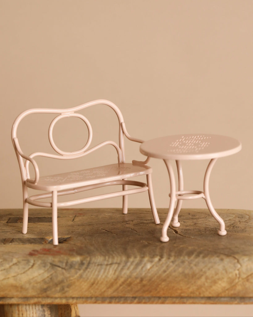 A small, light pink Maileg Patio Set—metal bench and matching round table with decorative cutouts—rests on a rustic wooden surface in front of a beige background.