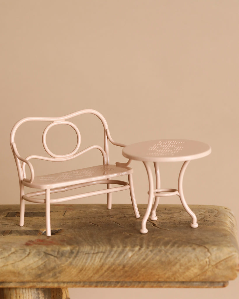 A small, pale pink metal table and chair set with intricate patterns sits on a rustic wooden surface against a plain beige backdrop. The Maileg Mouse Cafe Set furniture features curved designs, adding vintage charm ideal for an afternoon tea setting.