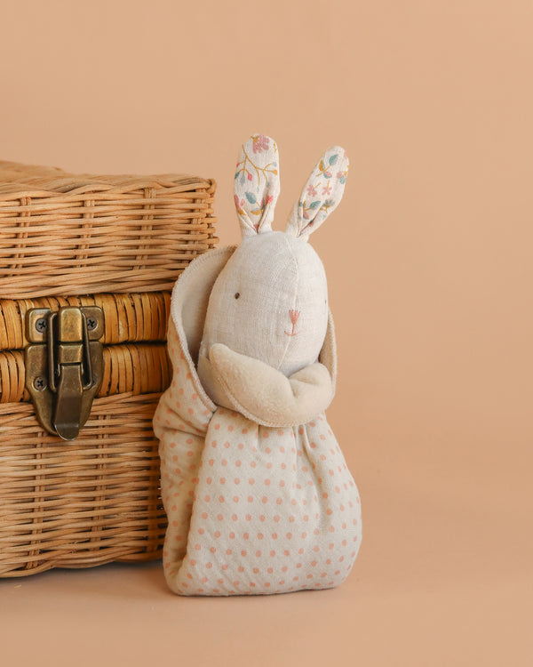 The Maileg Lullaby Friends Bunny, Mini - Nature features floral ears and a polka-dot body, sitting beside a closed wicker basket on a beige background—ideal as a cuddle cloth or charming newborn gift.