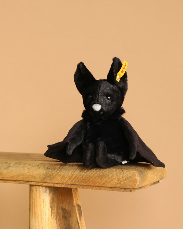 The Steiff Boris the Spooky Bat, a black plush toy with large ears and a white nose, sits on the edge of a wooden stool against a beige background, making an adorable Halloween accent.