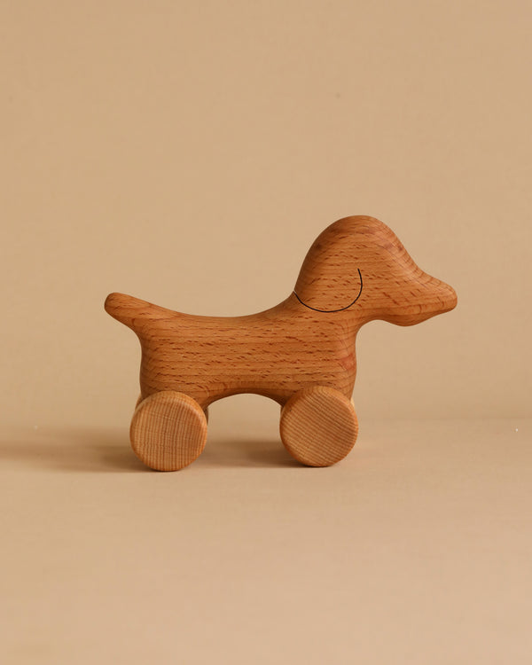 A Handmade Wooden Mini Dog Push Toy stands against a plain beige background. The toy boasts a smooth finish, simple carved details for the eyes and mouth, and four cylindrical wheels attached to its legs. The overall design is minimalist and natural.