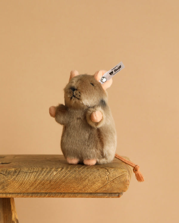 The Steiff Piff Mouse Plush Toy, 4 Inches, with a brown and beige body, pink ears, and a "Steiff" tag, stands upright on a wooden surface against a plain beige background.