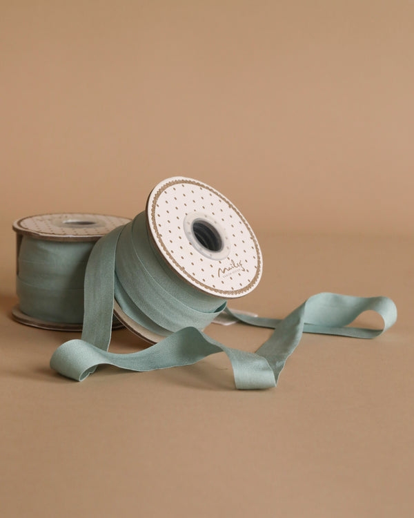 Two spools of Maileg Ribbon - Dusty Mint (10m), one upright and one lying down, are displayed on a neutral beige background. A length unrolls in front, ideal for adding an exclusive touch to decor or gift wrapping.