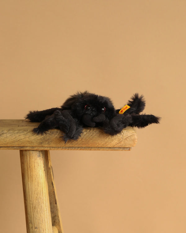 Steiff Cobweb the Spooky Spider, a black plush toy with red eyes, perches on a rustic wooden stool against a beige background—perfect for adding spooky Halloween charm to your decor.