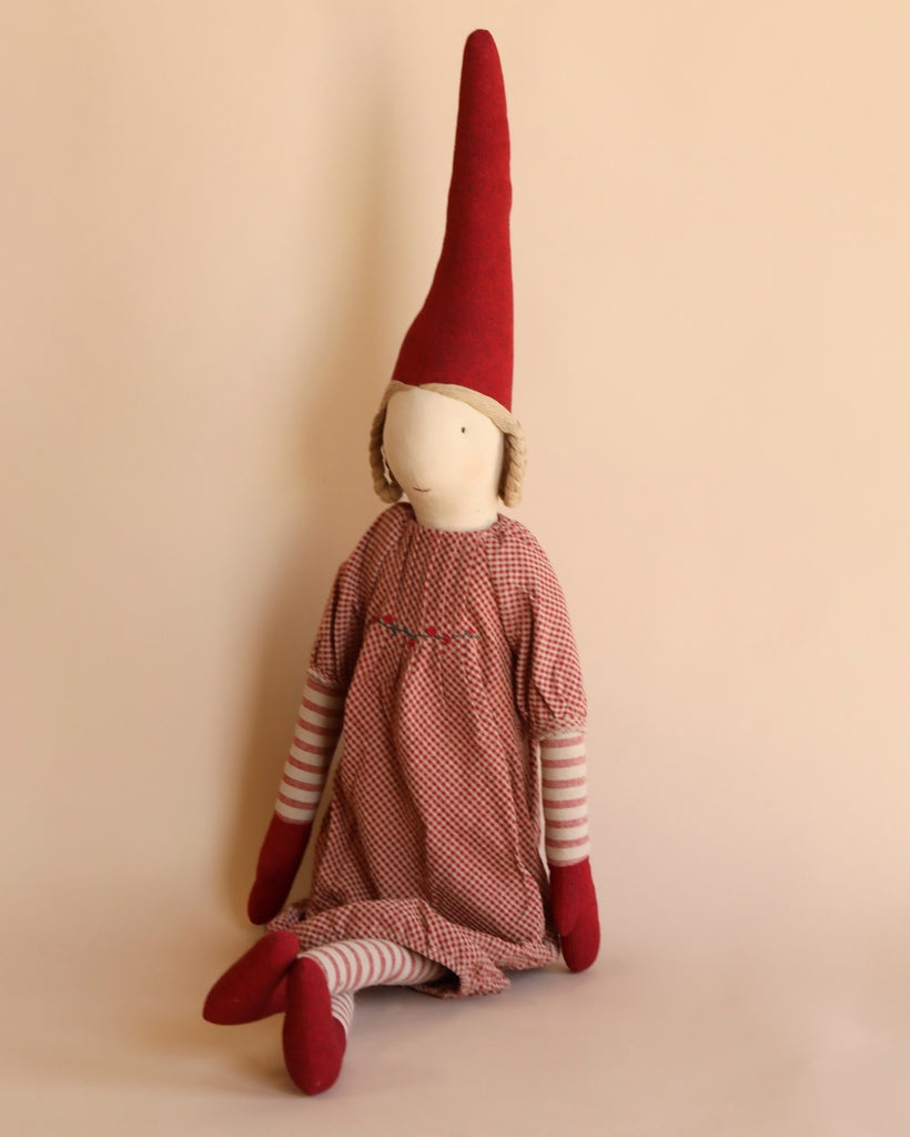 A Maileg Christmas Mega Pixy - (Size 6) doll with a red pointed hat, striped arms and legs, and an exclusive materials checkered dress in a seated position against a pale background.