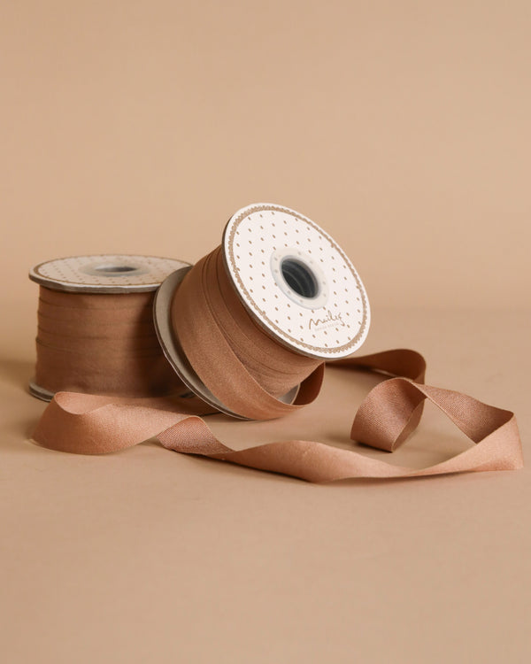 Two spools of Maileg Ribbon - Soft Brown (10m) rest on a beige background—one upright, one on its side with the ribbon unrolled—ideal for adding an exclusive touch to gift wrapping or special projects.