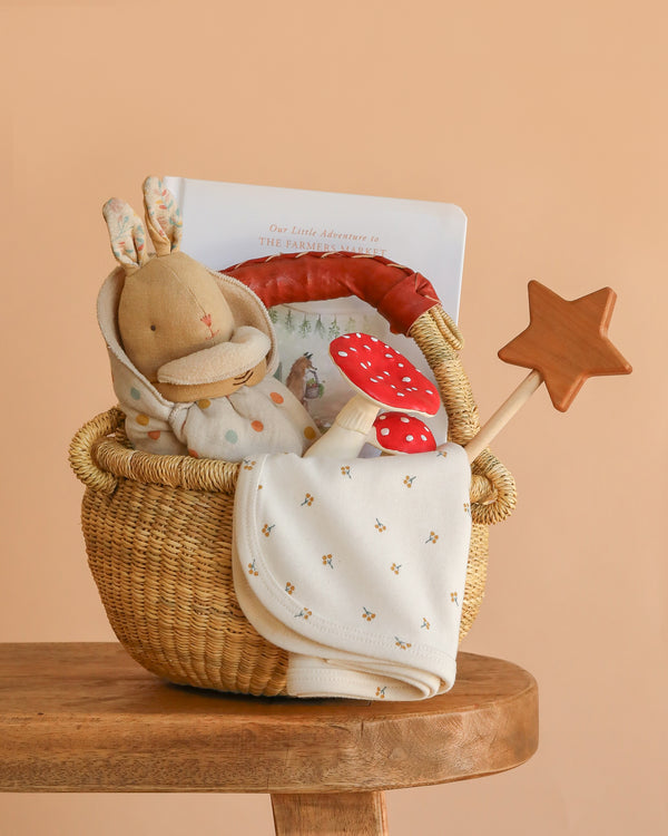 The Newborn Gift Basket Set, featuring a stuffed bunny, book, teething toy, star wooden wand, and organic baby blanket, is displayed on a wooden stool against a soft peach background.