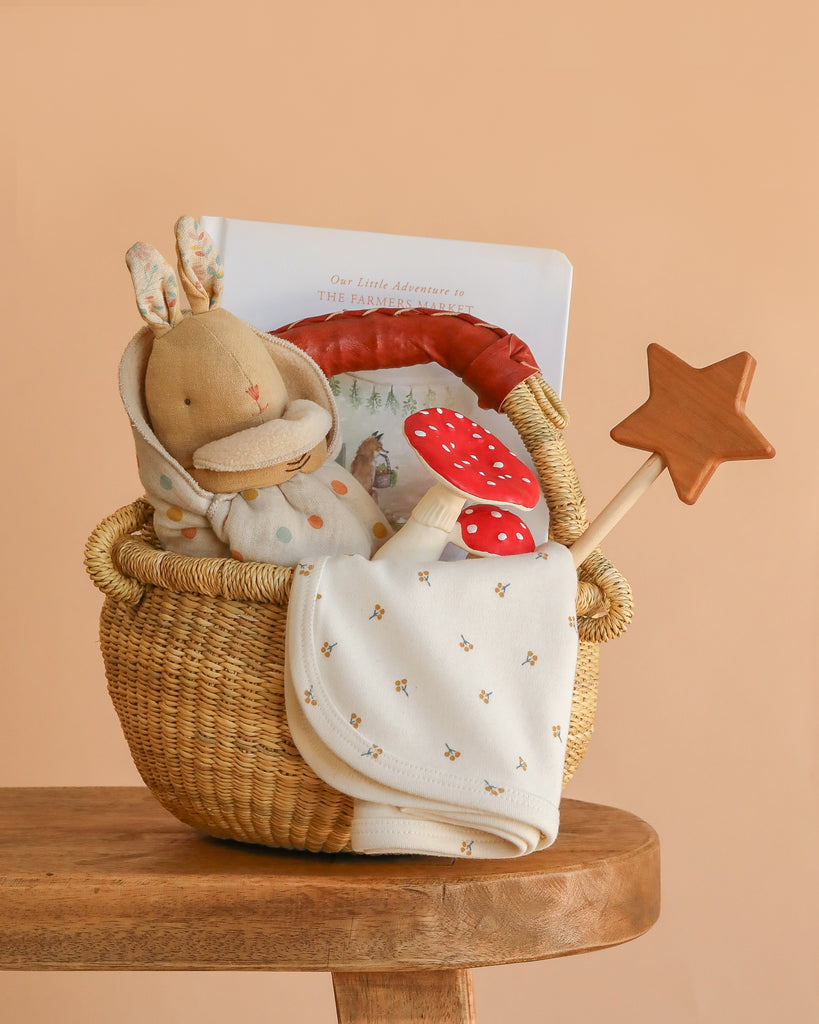 The Newborn Gift Basket Set, featuring a stuffed bunny, book, teething toy, star wooden wand, and organic baby blanket, is displayed on a wooden stool against a soft peach background.