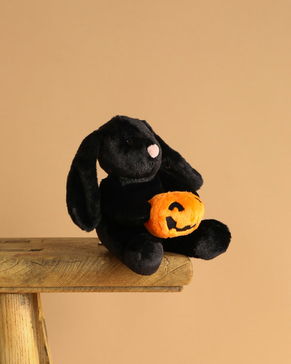 The Steiff Mr. Spice Hoppie Rabbit, a black bunny with floppy ears, sits on a wooden bench holding an orange jack-o'-lantern plush against a plain beige background.
