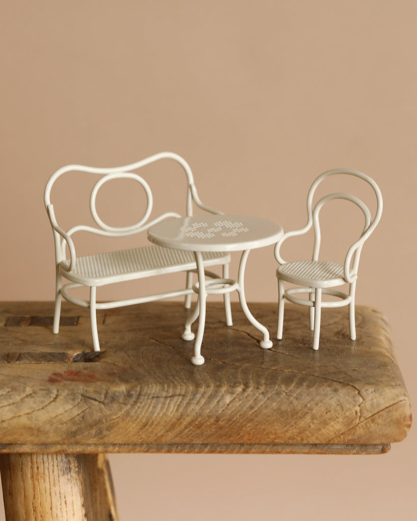 The Maileg Mouse Cafe Set, an off-white metal ensemble featuring a round table, bench, and chair, sits on a rustic wooden surface. Imagine afternoon sweets perfectly complementing this soft beige backdrop.