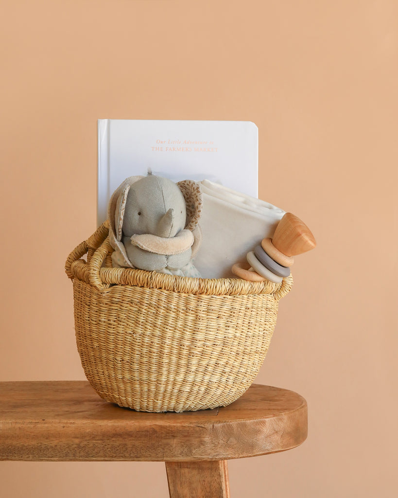 A Newborn Gift Basket Set sits on a wooden stool, featuring a gray stuffed elephant, Organic Baby Blanket, book, and Wooden Baby Rattle against a plain beige background.