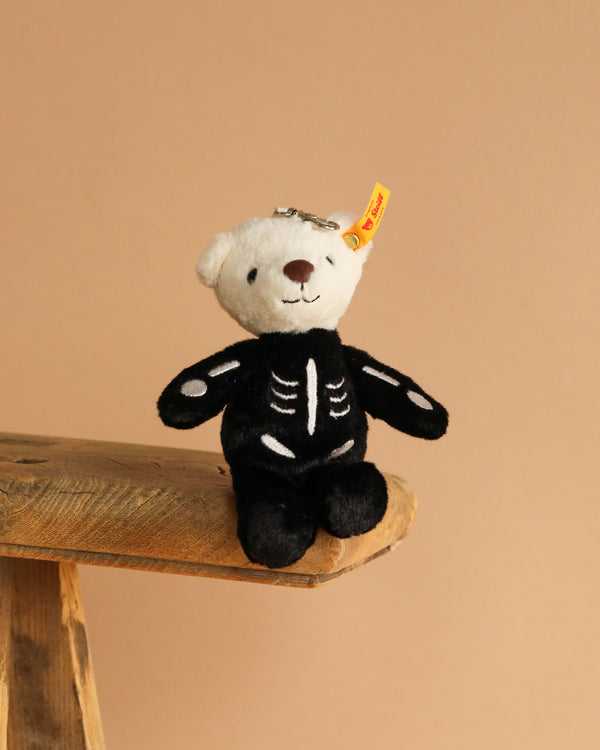 The Steiff Mr. Bones Teddy Bear Keychain, featuring a white head and black skeleton-themed body, sits on the edge of a wooden surface against a plain beige background.