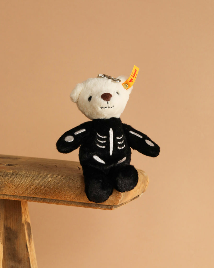 The Steiff Mr. Bones Teddy Bear Keychain, featuring a white head and black skeleton-themed body, sits on the edge of a wooden surface against a plain beige background.