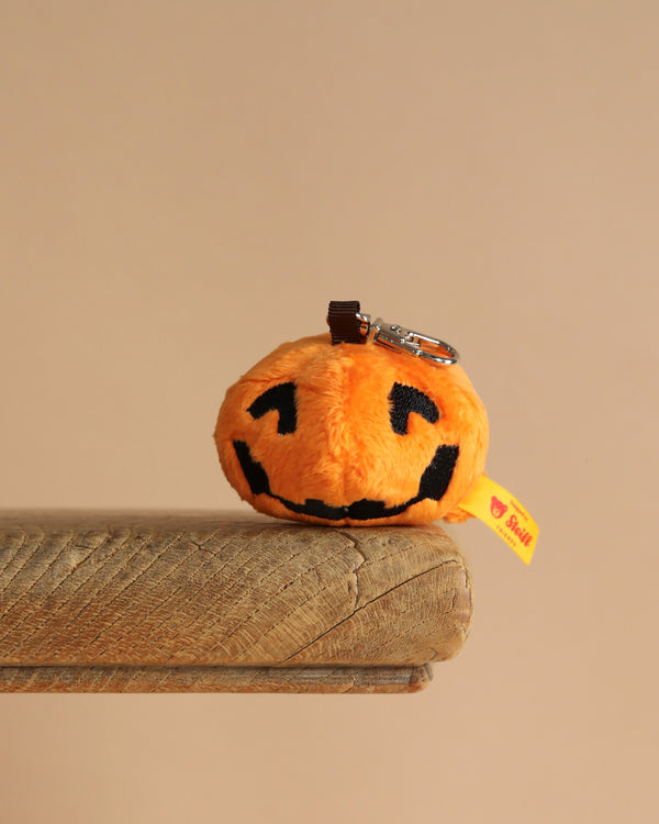 The Steiff Little Pumpkin Keychain, featuring a black jack-o'-lantern face, sits on the edge of a wooden surface against a plain beige background.