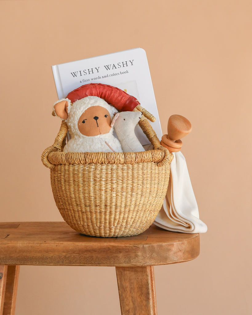 The Newborn Gift Basket Set, displayed on a wooden stool against a peach background, includes lamb and duck plush toys, a wooden baby rattle, an organic baby blanket, and the "WISHY WASHY" children's book.